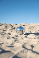 Vertical image of silver disco ball resting on sand dunes with a clear blue sky in background