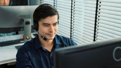 Young competent asian male call center agent working at his computer while simultaneously speaking...