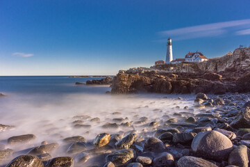 Fototapeta na wymiar Portland Head Lighthouse, Portland Maine. Lighthouse on cliff with keeper's quarters. Shot from ground level below cliffs. Bright sunshine. Long exposure water misting over rocks in foreground.
