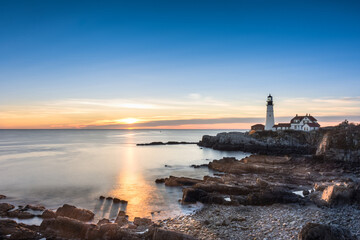 Sunrise at Portland Head Lighthouse, Portland, Maine. Looking across the water and rocks towards the lighthouse. Rays gleaming across the water shining on four rocks casting shadows