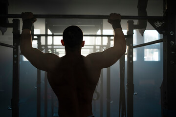 Fit shirtless man doing full chin-up exercise hanging on bar in sunny gym