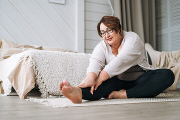 Middle aged plus size smiling brunette woman in home wear doing stretching in bedroom