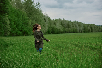 Naklejka na ściany i meble a beautiful woman in a long raincoat runs across a field in high grass in spring in cloudy weather