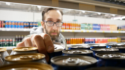 Close-up of many beer cans on a store shelf and a bearded man with glasses taking one