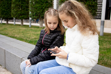 Portrait of two Charming smiling little girls chatting by video link sitting on a bench in the park. Technologies of modern communication, messages via the Internet using a mobile phone