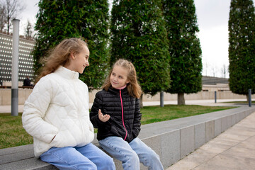 Two sister girls are sitting on a bench in the park, telling each other different stories or discussing something. Sincere and cordial relations between children.