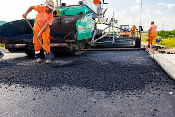 Workers with shovels realign hot asphalt. Spreader, tarmac road laying machine is in the background.