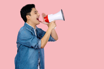 Happy young Asian man in blue shirt shouting announce into megaphone isolated on pink background in...