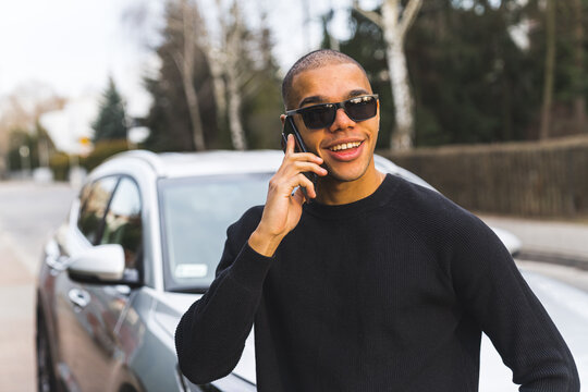 Positive smiling man in sunglasses standing in front of his car and calling someone with smartphone. Outdoor portrait. Communication concept. High quality photo