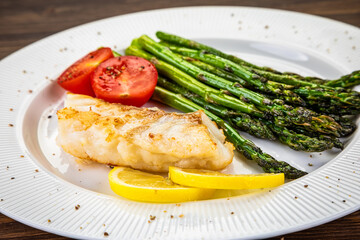 Fish dish - fried cod with green asparagus, lemon and fresh tomatoes on wooden table
