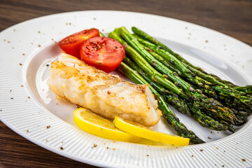 Fish dish - fried cod with green asparagus, lemon and fresh tomatoes on wooden table
