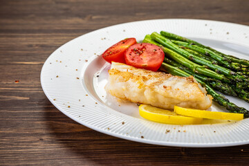 Fish dish - fried cod with green asparagus, lemon and fresh tomatoes on wooden table
