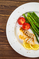 Fish dish - fried cod with green asparagus, lemon and fresh tomatoes on wooden table
