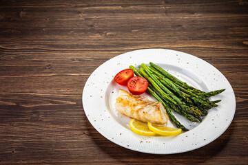 Fish dish - fried cod with green asparagus, lemon and fresh tomatoes on wooden table
