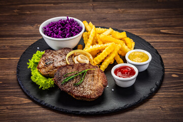 Fried beef sirloin with potatoes and vegetables on black stone plate on wooden table
