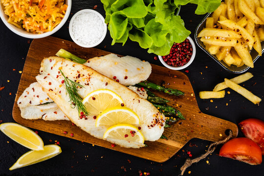 Fish dish - fried fish fillet with green asparagus and vegetables served on black stone plate on wooden table
