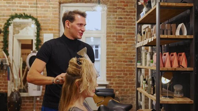 Professional hairdresser styling female client hair using hairdryer and round brush. A modern hair salon with mirror and rack in the background. High quality 4k footage