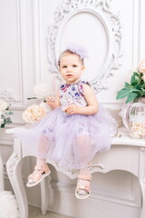 Fototapeta na wymiar Baby girl elegant dress. A one-year-old girl in a puffy dress and a cute bow poses against the backdrop of a bright room with a dressing table and flowers.