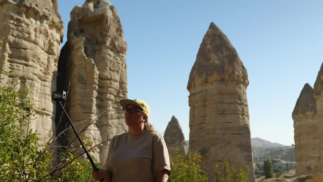 A blogger with glasses and a yellow baseball cap is recording a story with an action camera. In front of a sandstone rock in the shape of Phalos. The Valley of Love in Cappadocia. Holidays in Turkey