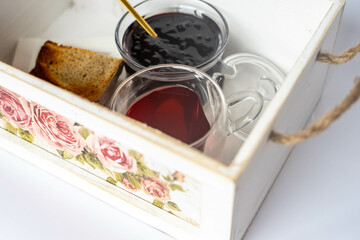 Obraz na płótnie Canvas Breakfast in a white wooden box, painted with pink roses. Cranberry jam, toast, red tea in a glass heart shaped mug.