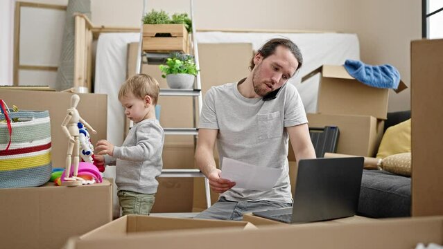 Father and son unpacking cardboard box using laptop talking on smartphone at new home