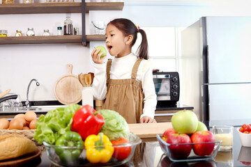 Asian cute girl kid eating green apple in modern kitchen with colorful fresh vegetables salad in a glass bowl and food ingredients on table counter kitchen, child and healthy food concept.