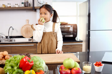 Asian cute girl kid eating green apple in modern kitchen with colorful fresh vegetables salad in a glass bowl and food ingredients on table counter kitchen, child and healthy food concept.