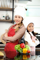 Portrait of happy smiling Asian mother daughter wear apron chef hat standing with arms crossed at kitchen with fresh vegetables salad food ingredient, cute chef family prepare cooking healthy food.