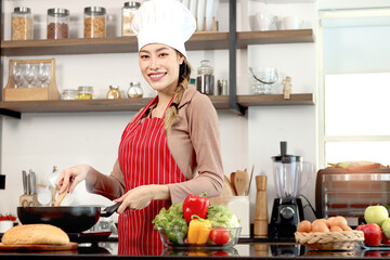 Happy smiling beautiful Asian woman wearing apron and chef hat, holding turner for cooking tasty fried food meal with pan at kitchen with fresh vegetable ingredient, female chef cooking healthy food.