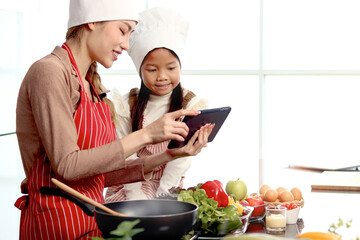 Happy Asian mother daughter wearing apron and chef hat at kitchen, mom and girl kid look at digital tablet for searching tasty healthy meal recipes on internet, cute chef family cooking food at home.