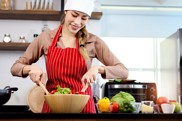 Happy smiling Asian beautiful woman wearing apron and chef hat, preparing fresh vegetables salad at kitchen, cute female chef preparing and cooking healthy food, vegan salad, diet, dieting concept.