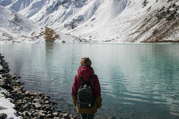 A medium shot photo of a woman standing alone with a backpack and looking at a high mountain lake