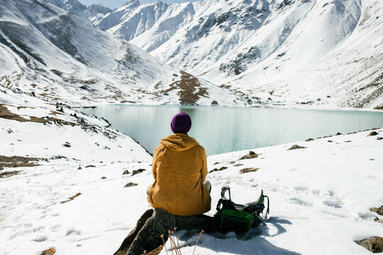 A wide shot photo of a man sitting alone with a backpack and looking at a high mountain lake