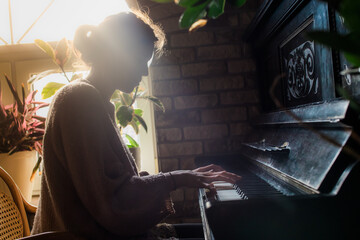 woman playing the old rustic piano with a beautiful ray of sunlight from behind, side view,...