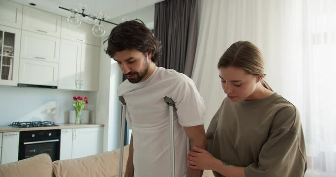 Wife is helping her handsome husband with broken leg to move along the room, slow motion