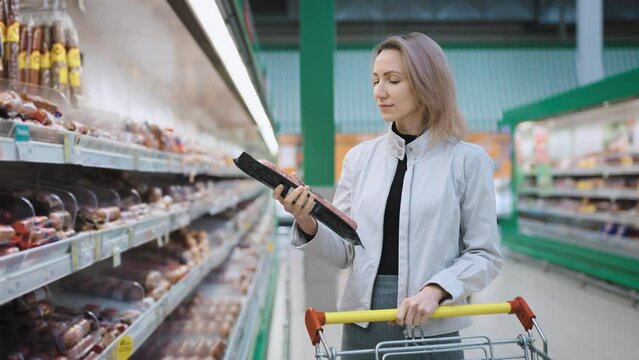 A blonde woman with a grocery cart stands near a shelf with sausages in a super market