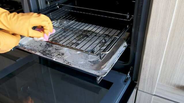 A man in protective gloves uses a kitchen sponge to clean the oven grate from burnt-on dirt. Up close