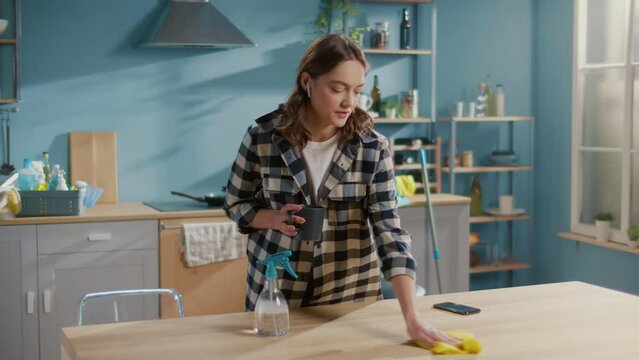 A Young Female Housewife in Headphones is Talking to Someone While Cleaning the Table and Drinking Coffee. Multitasking Cleaning Routines.Efficient Work-from-home Setups.Home Office Organization Tips.