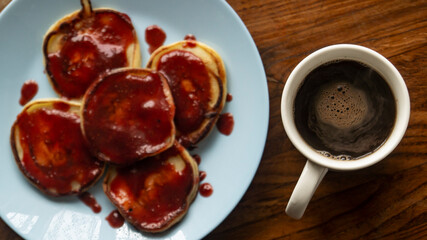 Pancakes on a blue plate. Pancakes with red sauce. Strawberry jam and pancake on a plate. Black coffee in a white cup and a plate with pancakes on a wooden table. Breakfast with coffee