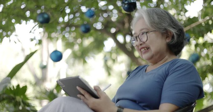 Asian elderly woman happily sitting in her backyard reading a book or shopping online using a tablet. technology for the elderly. online communication with family
