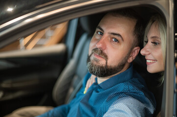 Happy married couple testing new car in car showroom. 