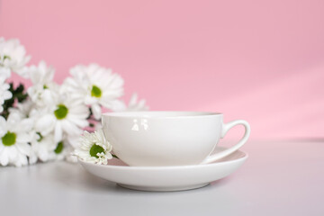 Obraz na płótnie Canvas A white cup with a plate stands on a pink background with white chrysanthemum flowers