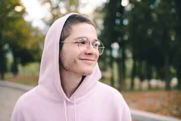 Smiling cute teenager boy 19-20 year old wear glasses and hood over nature background outdoor close up. Happy male kid student.