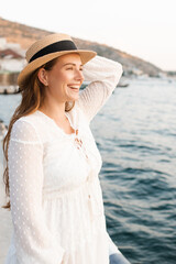 Laughing young adult woman wear straw hat and white stylish rustic dress over sea outdoor. Summer vacation season.