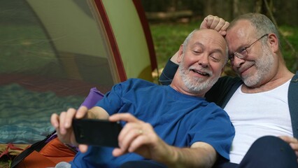 Closeup of two gay men in front of tent with pride flag at campsite taking selfie with phone.