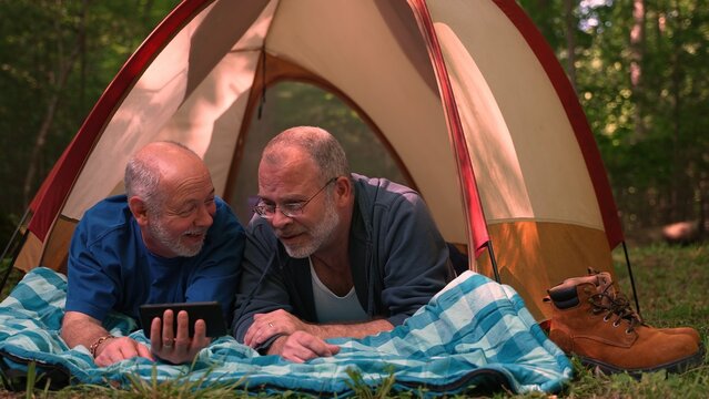 Two gay men with pride flag and tent in front of campfire talking to smartphone on video chat or taking selfie.