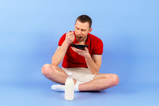 Young handsome healthy man in a red polo shirt is eating cereal or breakfast with milk. Isolated on blue background. full length.