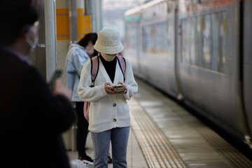 駅のホームで電車を待ちながらスマートフォンを弄る若い女性の姿 - obrazy, fototapety, plakaty