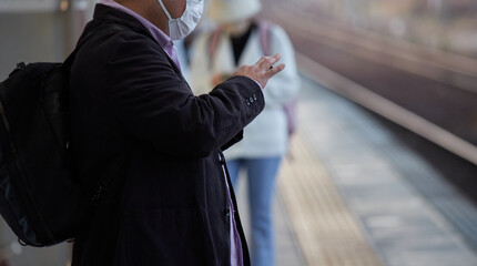 駅のホームでマスクをしてスマートフォンを弄る電車待つ男性サラリーマンの姿 - obrazy, fototapety, plakaty