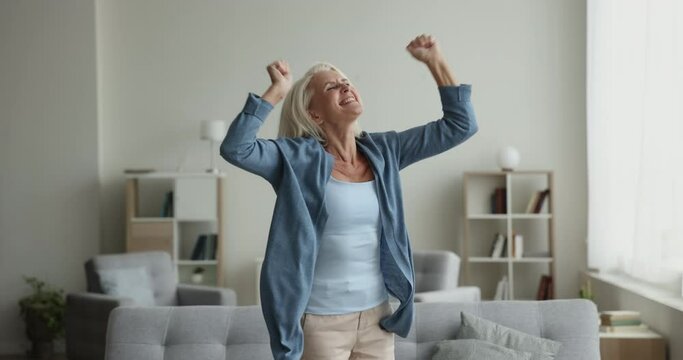 Untroubled mature woman dance in living room, have fun at home listens song, moves to favourite track feels overjoyed, celebrate good news, relish carefree retired life looks happy. Vitality, wellness